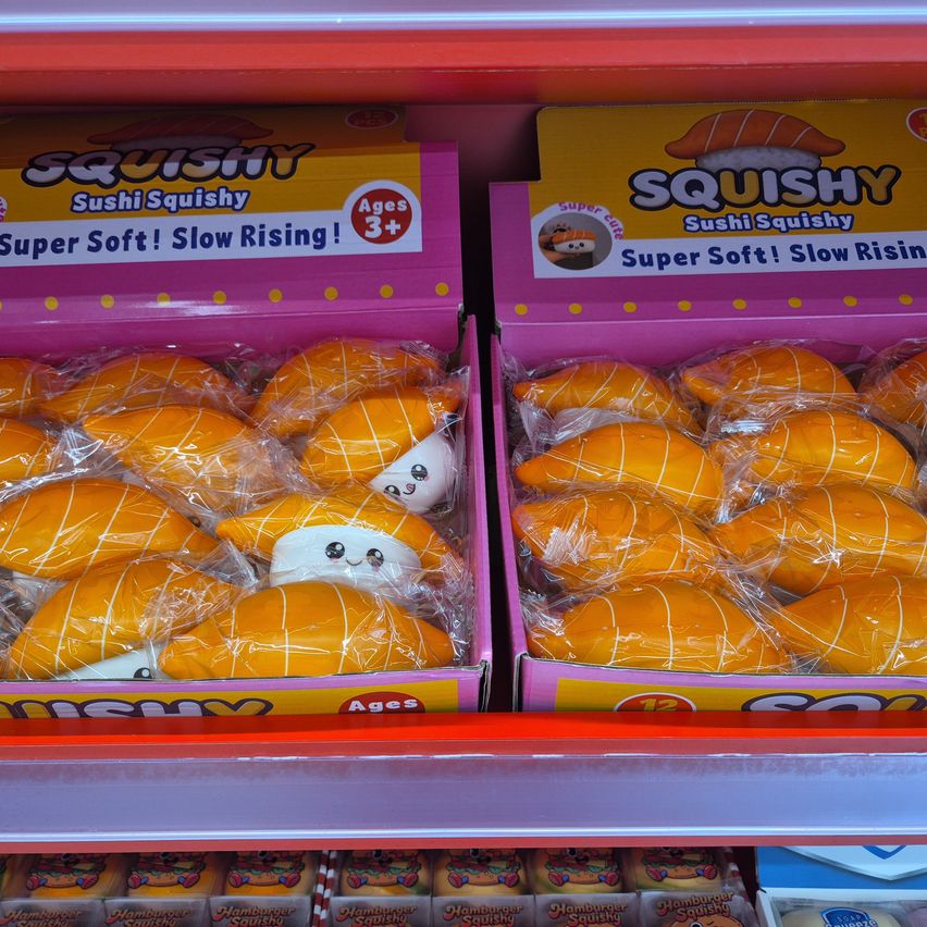 Zwei Boxen Squishy Sushi Squishy Spielzeuge stehen in einem Ladenregal. Jede Box enthält orangefarbene, sushiförmige Spielzeuge, die in Plastik verpackt sind. Die Schachteln haben farbenfrohe Etiketten mit den Texten 'Super Soft! Slow Rising!' und 'Alter 3+' darauf.