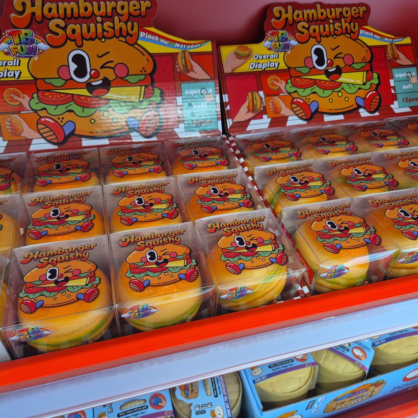 Ein Ladenregal zeigt zahlreiche durchsichtige Plastikverpackungen von Hamburger Squishy-Spielzeug, jedes mit einer fröhlichen Cartoon-Hamburger-Figur mit Augen und einem lächelnden Gesicht.