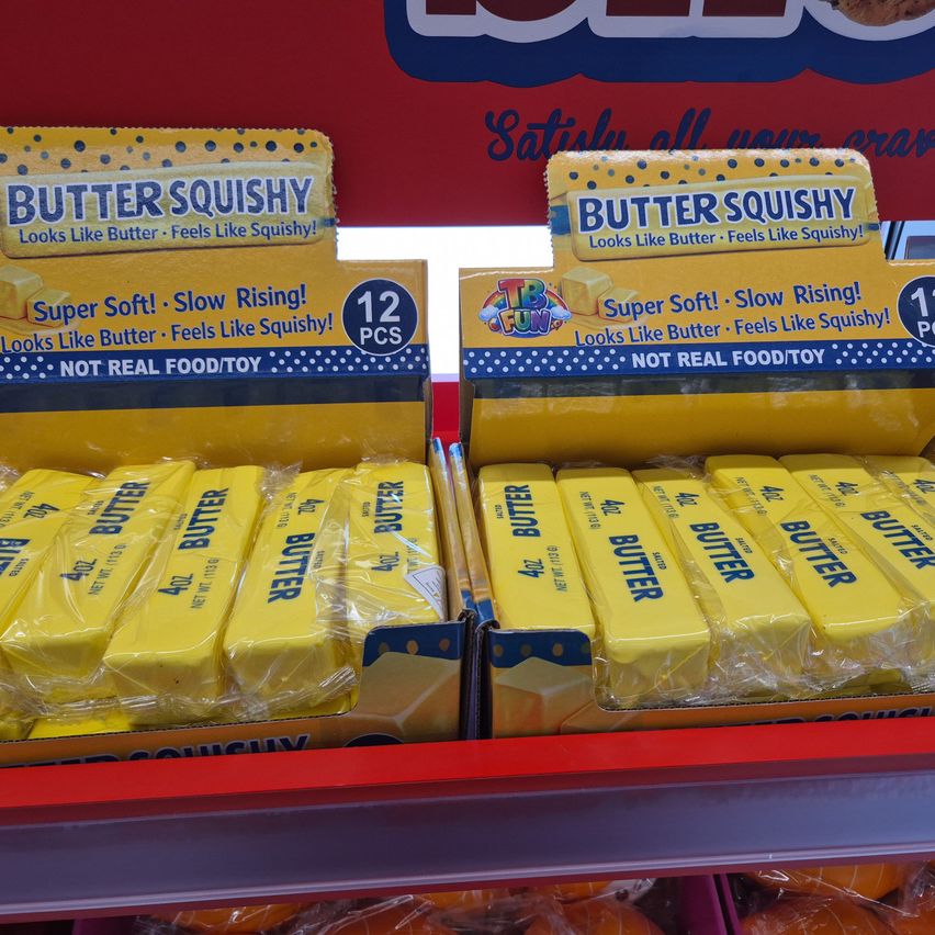 Zwei Schachteln mit Butter Squishy sind auf einem Regal ausgestellt. Jede Schachtel enthält 12 Stück, und das Produkt ist in Plastik verpackt. Die Schachteln haben einen gelben Hintergrund mit blauen Punkten und dem Text 'Superweich! Langsam aufgehend! Sieht aus wie Butter Führt sich wie Squishy an!'. Das Produkt ist keine echte Nahrung oder ein Spielzeug.