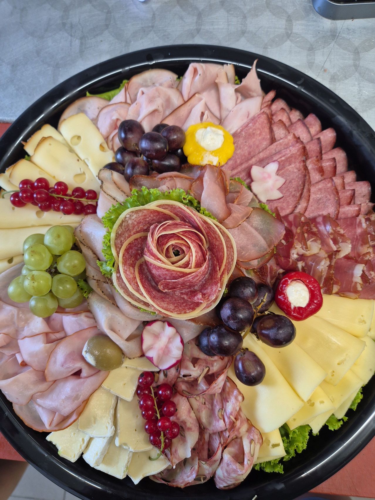 Ein schwarzer Teller gefüllt mit einer Auswahl an Fleisch, Käse, Trauben und Oliven, mit einer dekorativen Fleischrose in der Mitte.