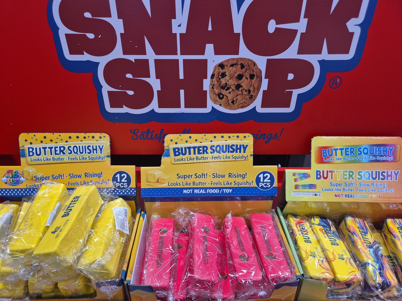 A display of Butter Squishy toys in various colors and packaging, with a sign reading 'Snack Shop' in the background.