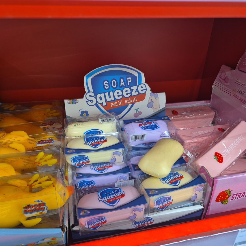 A store shelf displays various soap products, including yellow and pink soap squeeze toys, with a sign that reads 'SOAP SQUEEZE Pull it! Rub it!'.
