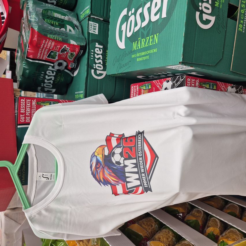 A white t-shirt with a soccer design and the text 'WM 26' is displayed on a hanger. Behind it, several boxes of Gösser beer are stacked.