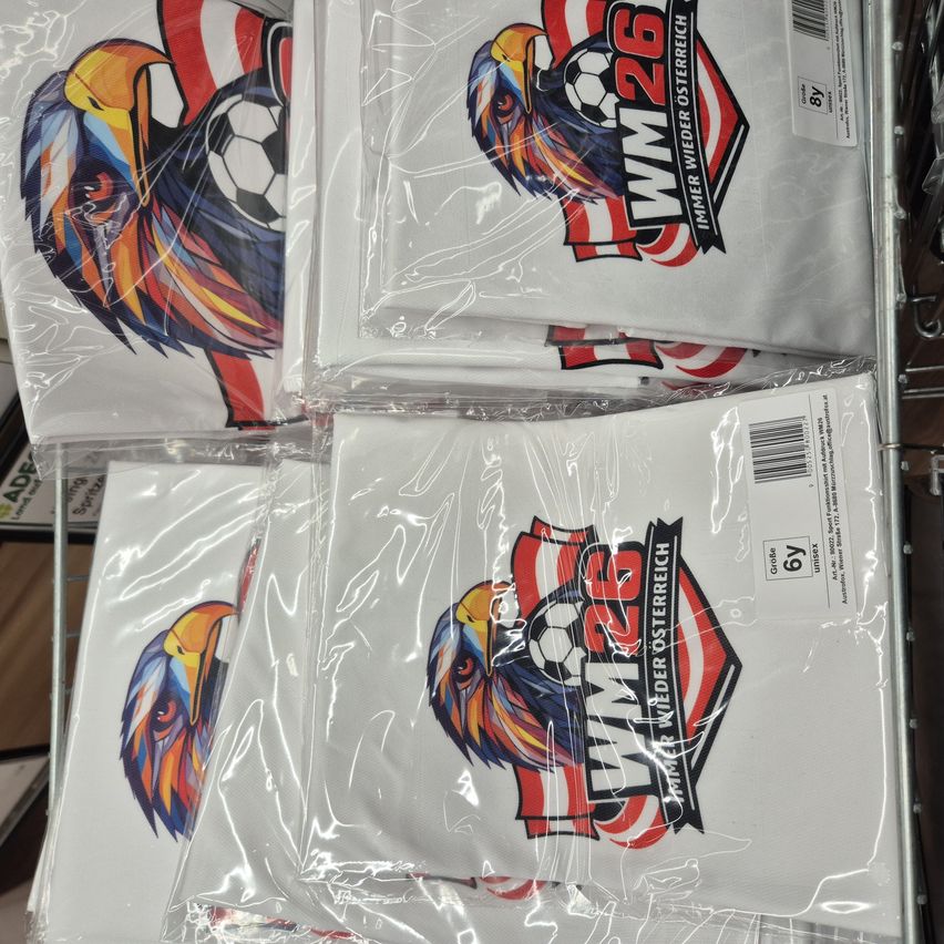 Several white soccer jerseys with the WM26 logo and a colorful eagle are stacked and sealed in plastic. Each jersey has a barcode label.