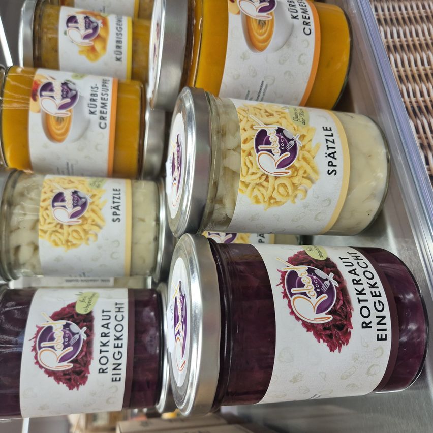 Multiple jars of assorted foods, including sauerkraut, pasta, and curd cheese, are displayed on a metal surface. The jars have white labels with illustrations and text.