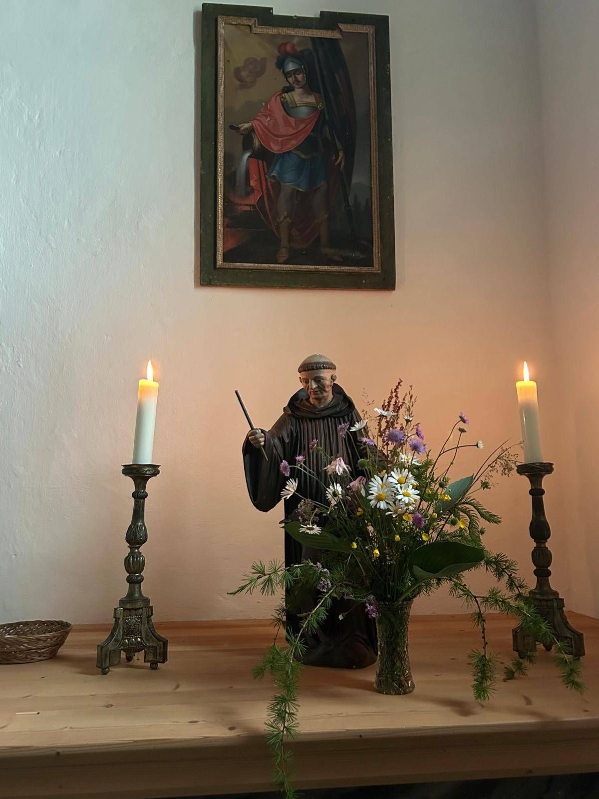 Eine Statue des Heiligen Franz von Assisi steht vor einem Blumenstrauß. Die Statue hält einen Stock und trägt einen Mantel. Zwei brennende Kerzen flankieren die Statue. Ein Gemälde hängt über der Szene.