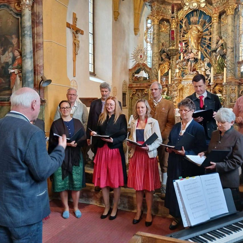 Eine Gruppe formell gekleideter Menschen steht in einer Kirche, hält Bücher und singt. Ein Klavier und Notenblätter stehen davor. Ein Kreuz und eine Statue sind dahinter.