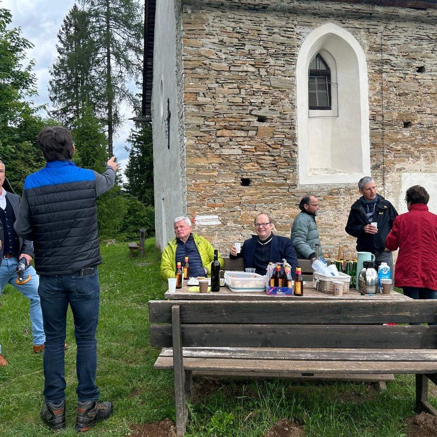 Eine Gruppe von Menschen versammelt sich draußen in der Nähe eines alten Gebäudes mit Steinmauern. Ein Mann steht und gestikuliert, während andere an einem Tisch mit Flaschen und Essen sitzen.