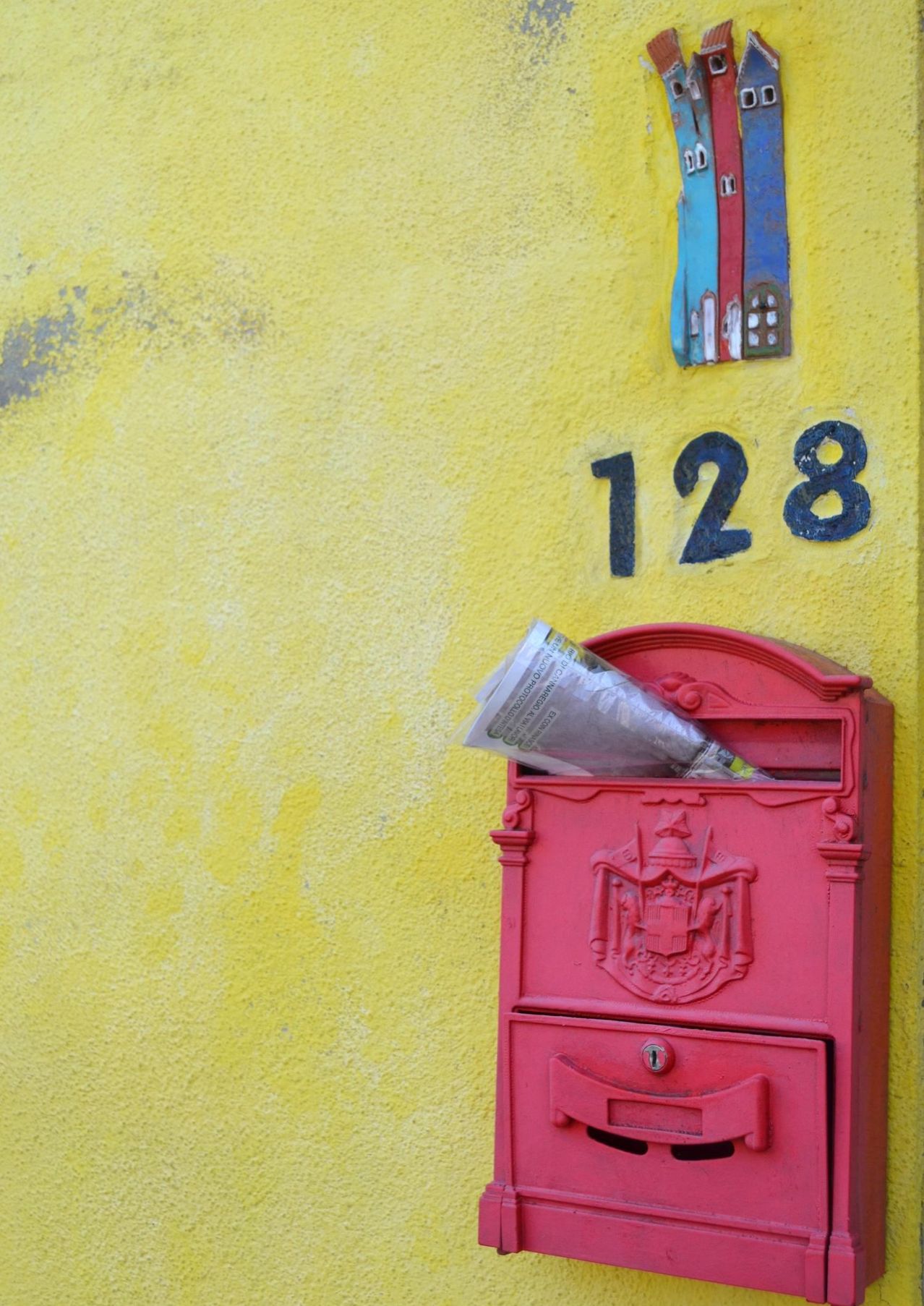 Eine gelbe Wand mit einem roten Briefkasten, die Nummer 128 darüber gemalt und eine Zeitung darin.