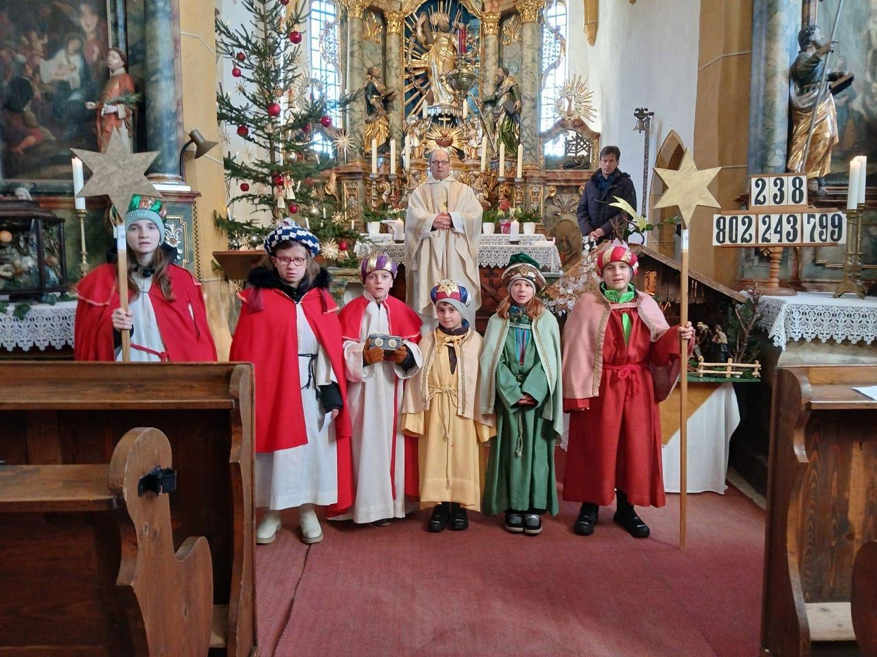 Eine Gruppe von Kindern in traditionellen Kostümen steht in einer Kirche, möglicherweise während einer Weihnachtsfeier. Ein Priester steht hinter einem Altar mit einem Weihnachtsbaum und Dekorationen.