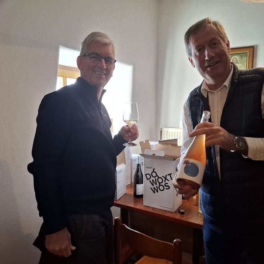 Zwei Männer stehen in einem Raum und lächeln. Einer hält ein Weinglas, der andere eine Flasche Wein. Hinter ihnen steht ein Tisch mit Weinkartons, einer ist mit 'Do Woxt Wos' beschriftet. Ein Bilderrahmen hängt an der Wand.