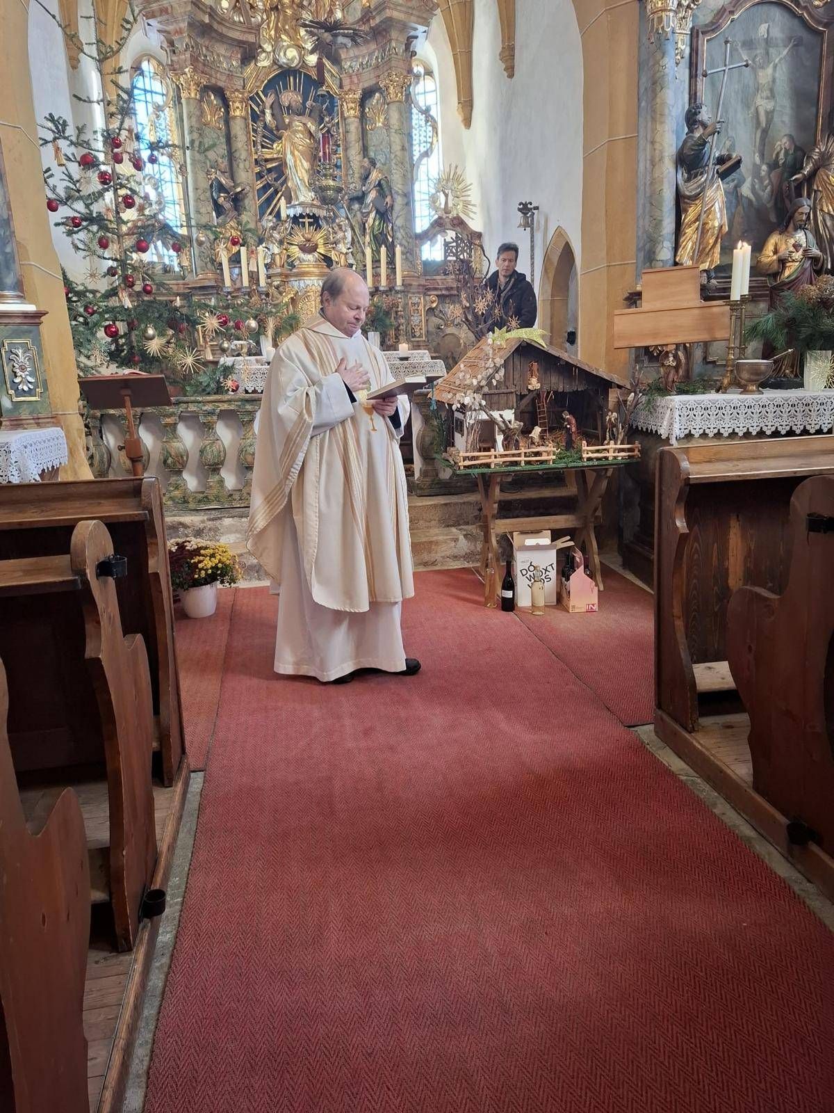 Ein Priester steht in einer Kirche und liest ein Buch. Hinter ihm ist eine Krippe auf einem Tisch ausgestellt. Ein Weihnachtsbaum und Kerzen schmücken den Altar. Eine weitere Person steht in der Nähe des Altars.