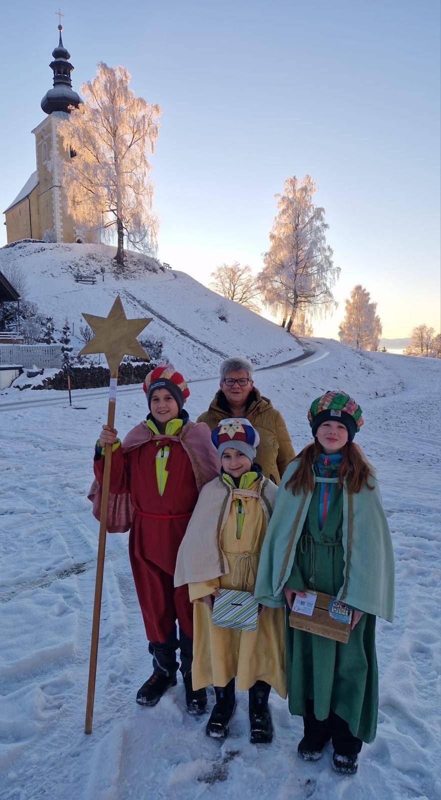 Drei Kinder in traditionellen Kostümen posieren für ein Foto im Schnee. Eines hält einen sternenbesetzten Stab. Eine Frau steht hinter ihnen, und ein Haus ist im Hintergrund sichtbar.