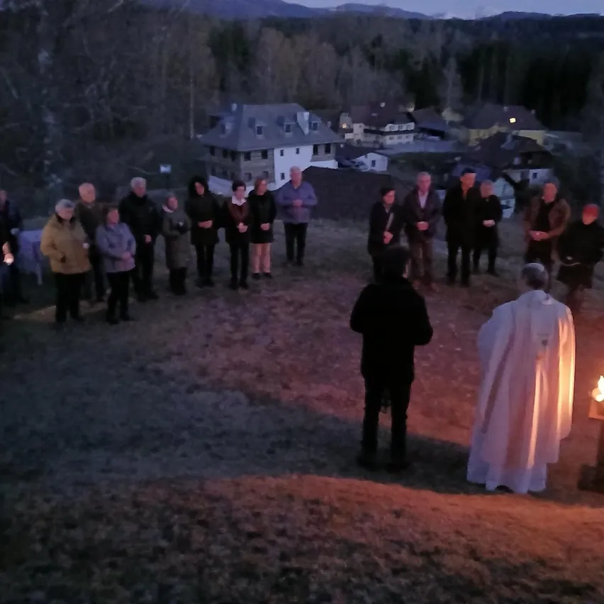 Eine Gruppe versammelt sich für eine abendliche Zeremonie mit einem Priester vor einer brennenden Kerze, umgeben von Bäumen und Gebäuden in der Ferne.