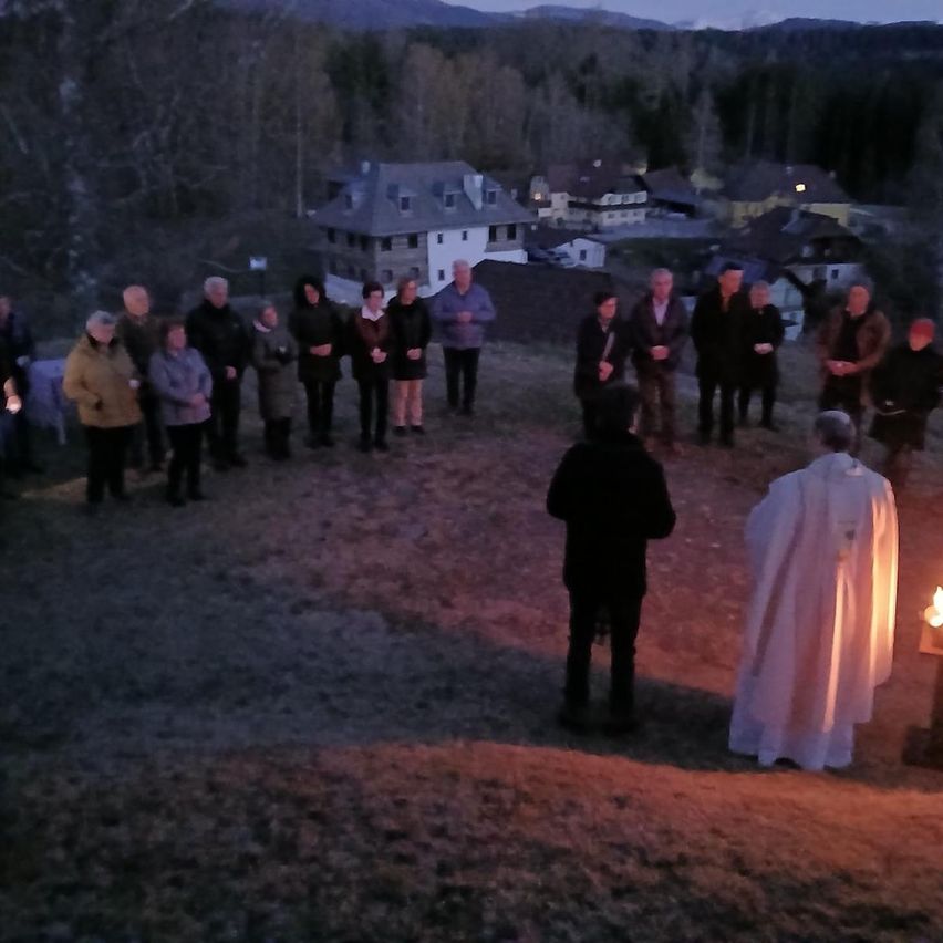 Eine Gruppe versammelt sich für eine abendliche Zeremonie mit einem Priester vor einer brennenden Kerze, umgeben von Bäumen und Gebäuden in der Ferne.