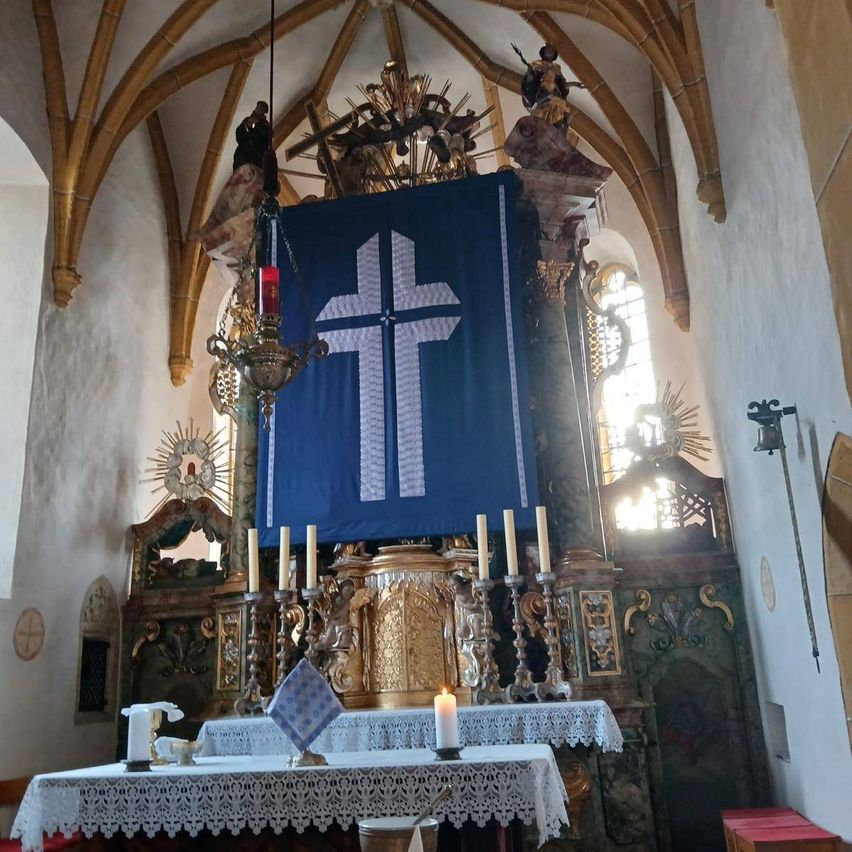 Ein Kirchenaltar mit einem blauen Banner, das ein weißes Kreuz zeigt. Vorne sind Kerzen aufgestellt, und eine einzelne Kerze ist angezündet. Der Altar hat eine weiße Tischdecke mit komplizierter Spitze.