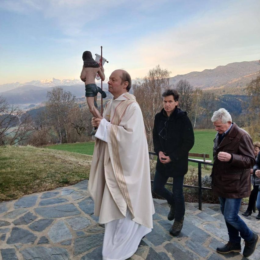 Ein Priester hält eine Statue und geht auf einem Pfad mit zwei Männern. Dahinter befindet sich eine Wiese mit einer Bank und Bergen in der Ferne.