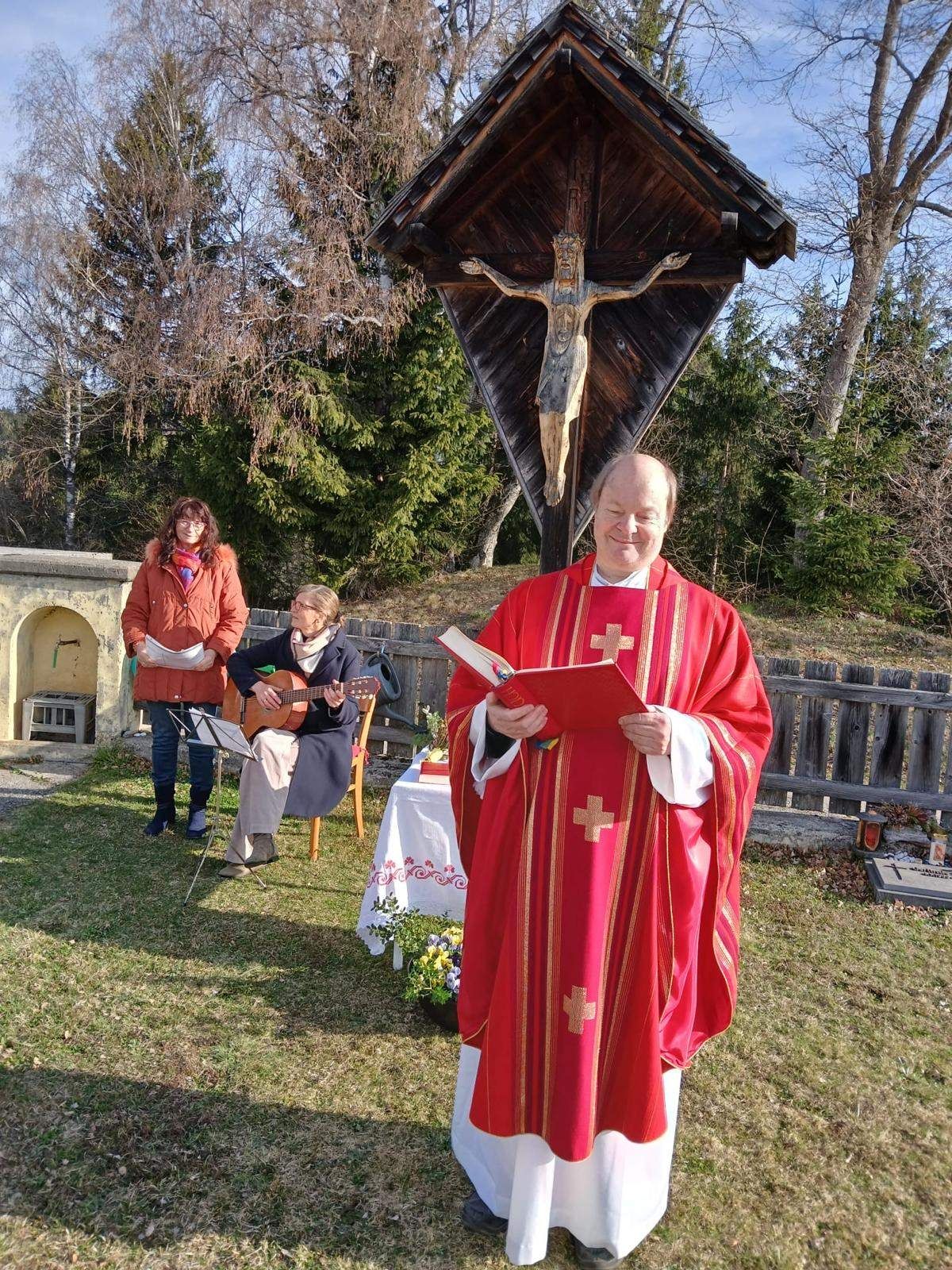 Ein Priester liest ein Buch vor einem Kreuz. Eine Frau spielt neben ihm Gitarre. Eine andere Frau steht mit einem Notenständer hinter ihnen.