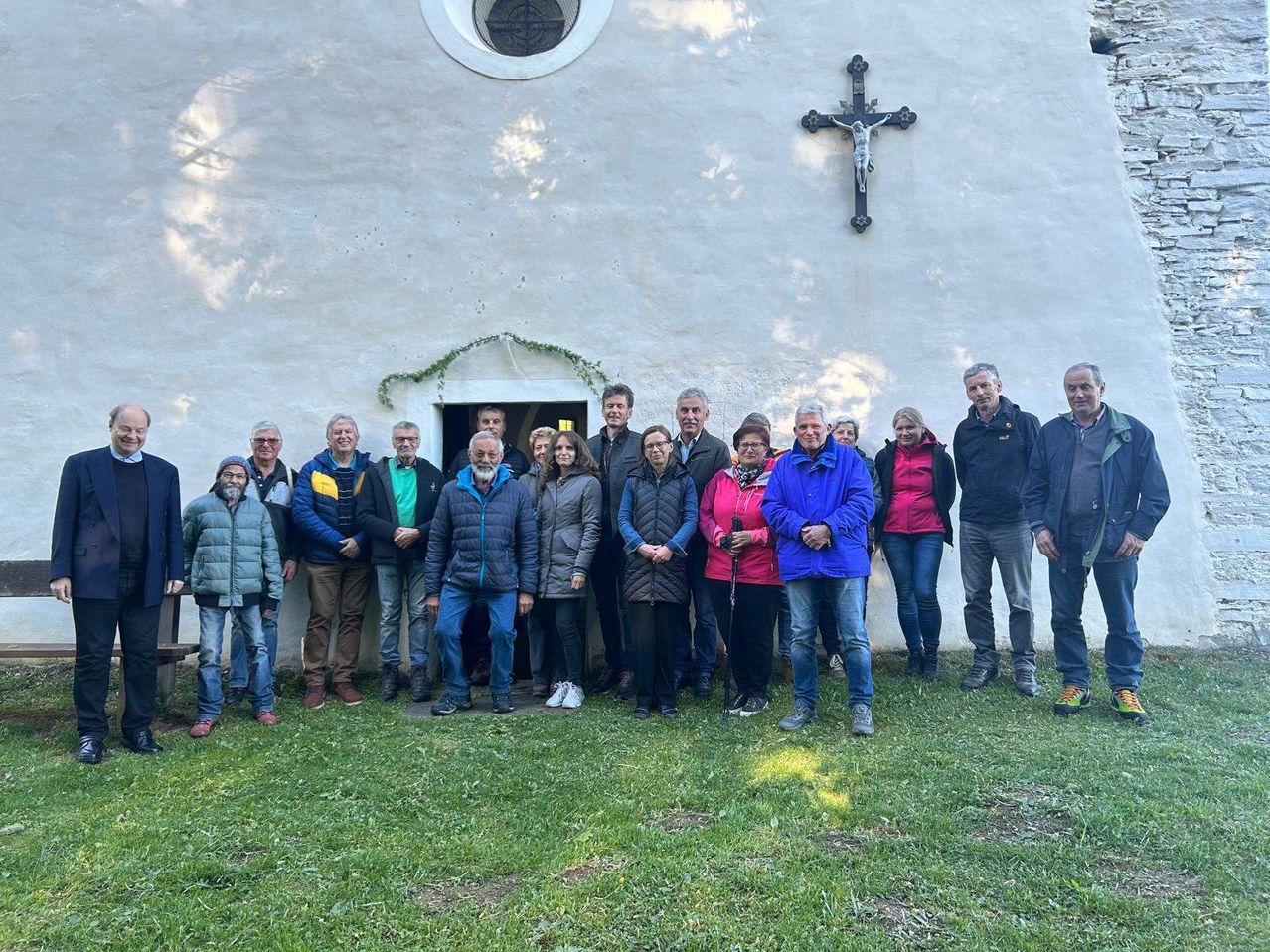 A group of people stand in front of a white church with a cross on the wall. They are wearing various jackets and pants. Some of them are holding sticks.