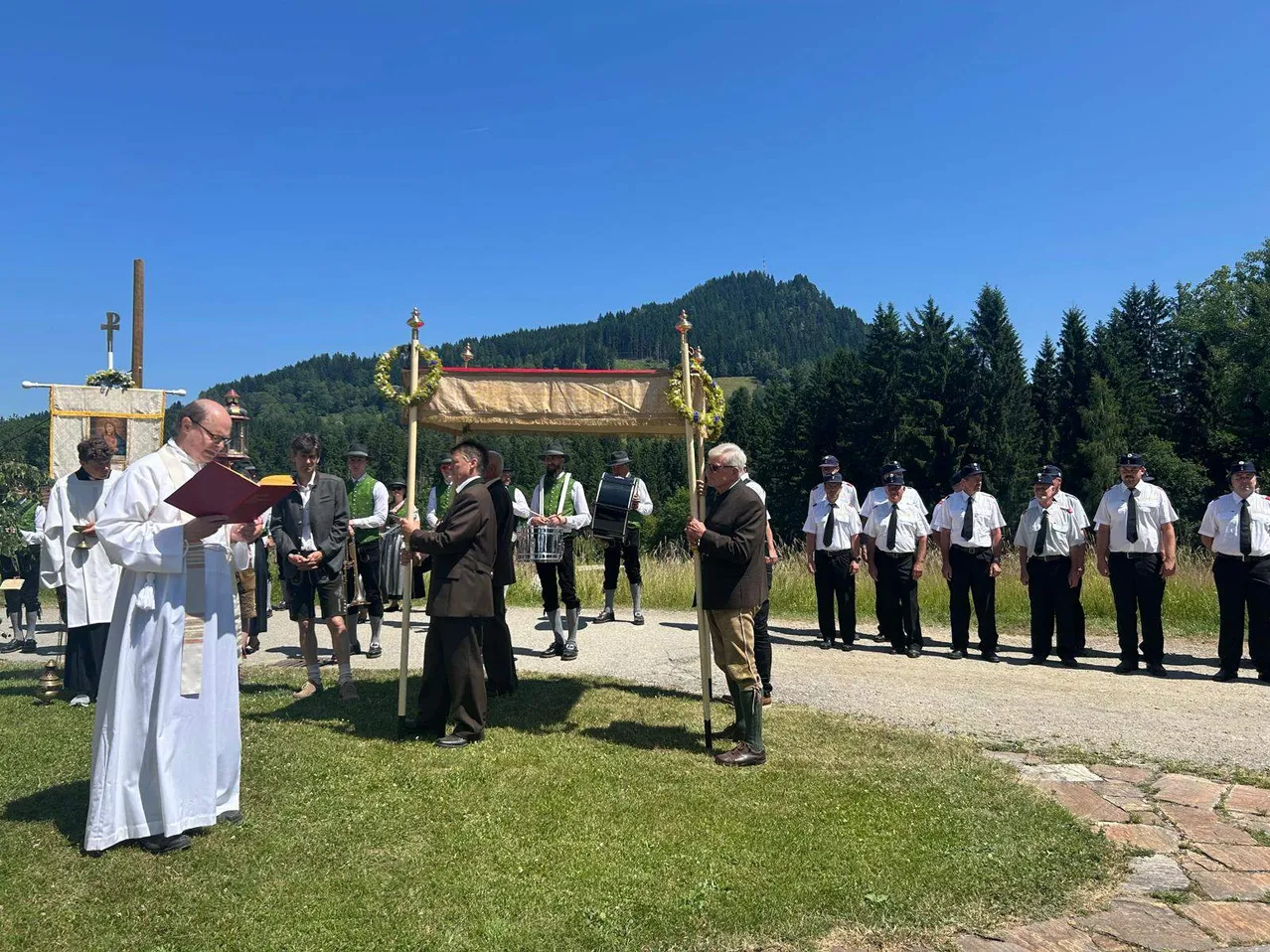 Ein Priester führt eine Zeremonie im Freien durch, wobei die Teilnehmer in traditioneller Kleidung stehen und Berge im Hintergrund zu sehen sind.