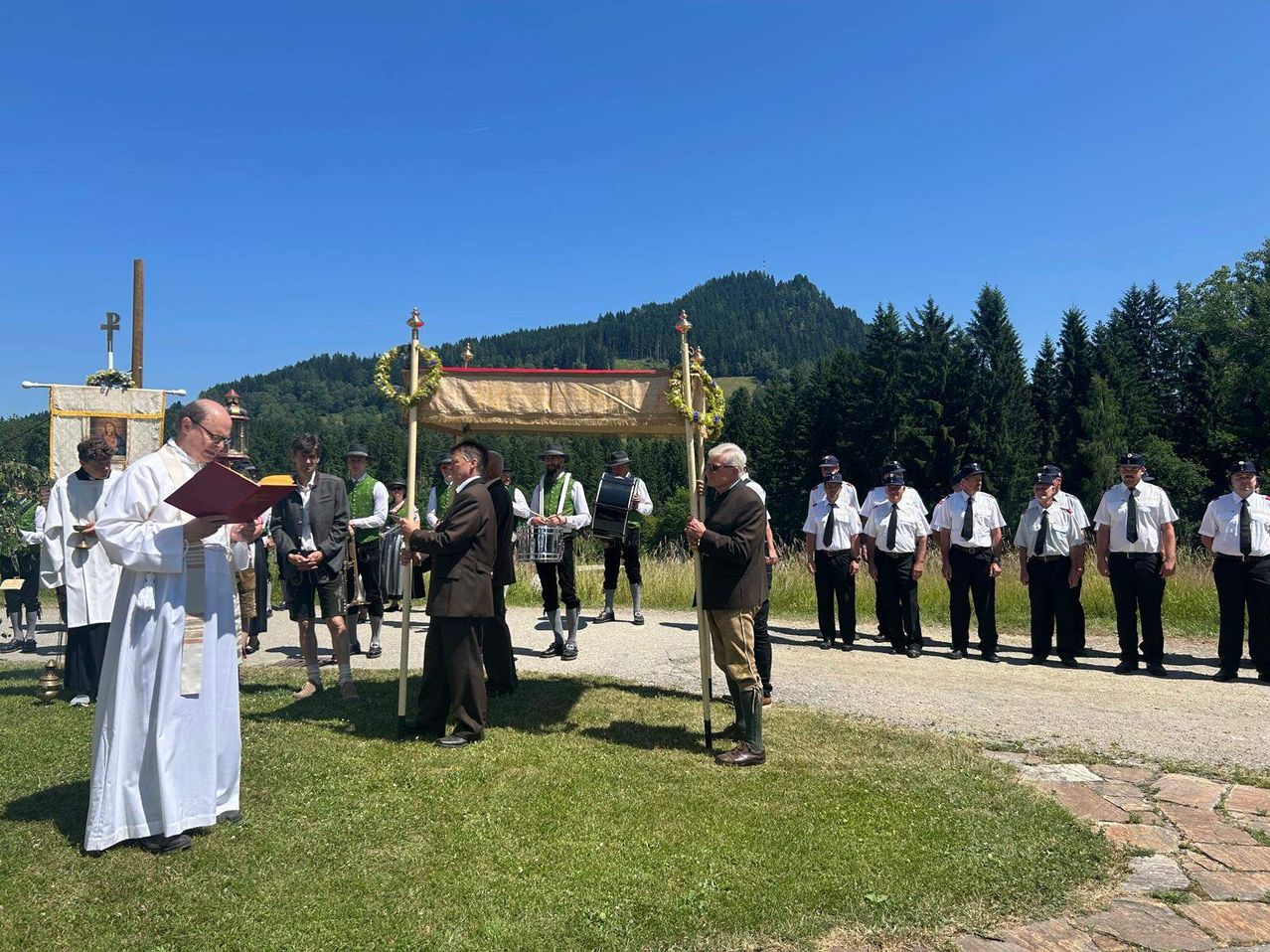 A priest conducts a ceremony in an outdoor setting, with participants in traditional attire and mountains in the background.