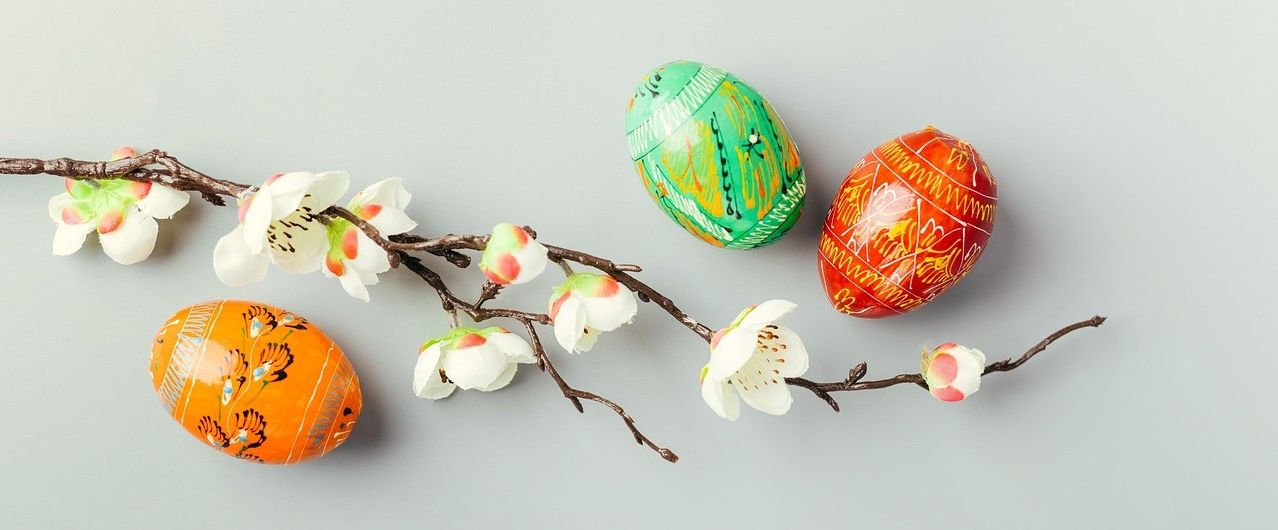 Decorated Easter eggs rest on a branch with white flowers on a gray background. The eggs are painted in various colors and patterns.
