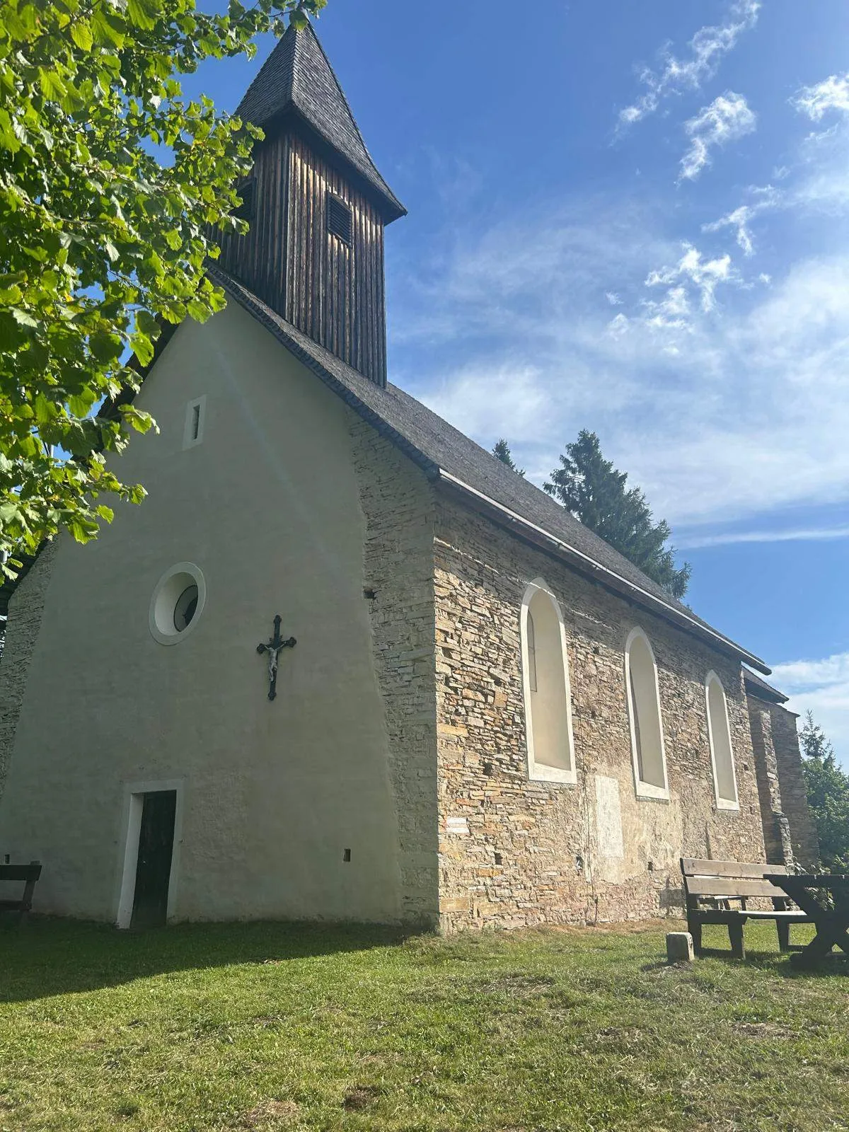 Eine kleine Steinkirche mit einem hölzernen Turm und einem Kreuz an der Fassade, umgeben von Bäumen und einer Bank.