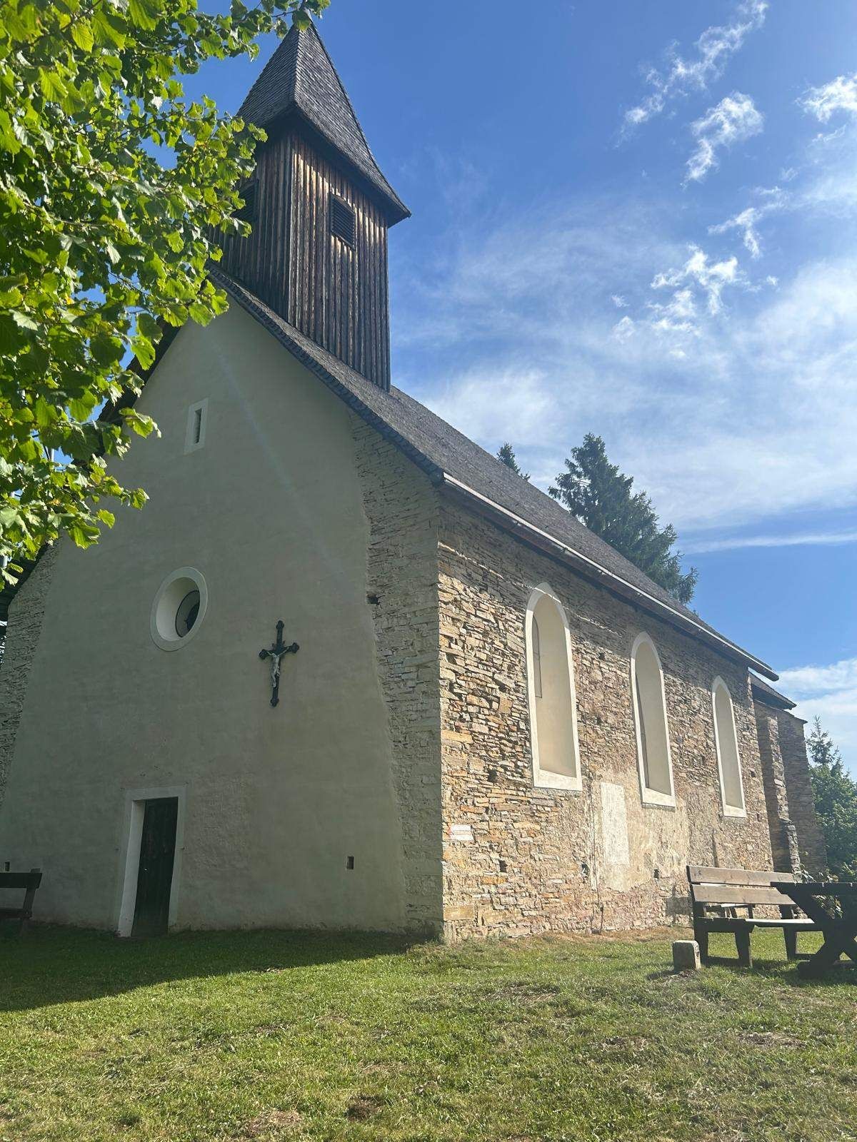 A small, stone church with a wooden tower and a cross on its facade, surrounded by trees and a bench.