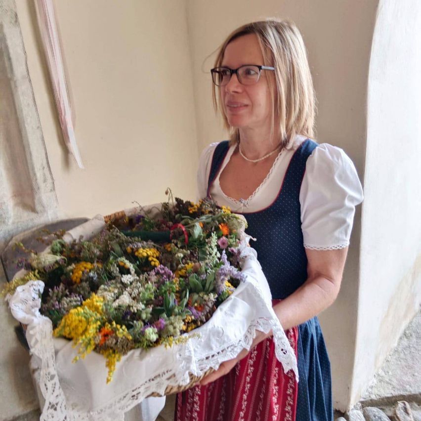 Eine Frau in traditioneller bayerischer Tracht hält einen Korb voller verschiedener Kräuter und Blumen. Sie steht an einer Steinmauer.