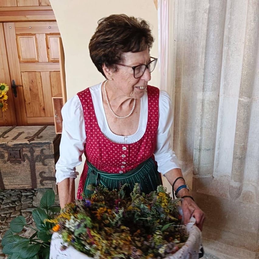 Eine ältere Frau in einem traditionellen deutschen Kleid und mit Brille hält einen Korb voller getrockneter Blumen. Sie steht vor einer Steinmauer.