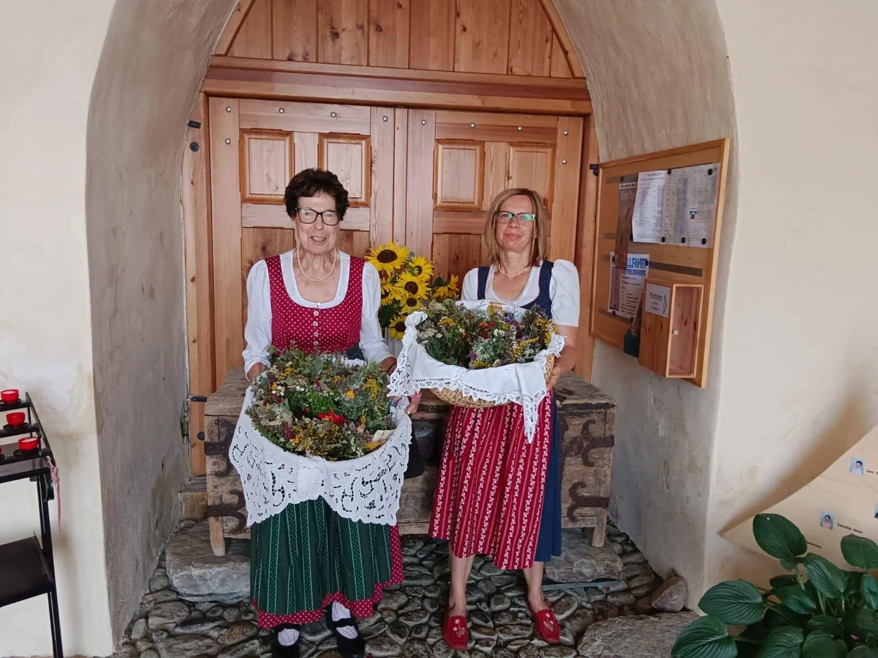 Zwei Frauen in traditioneller Kleidung stehen vor einer Holztür, jede hält einen Korb mit Blumen. Die Frau auf der linken Seite trägt ein rotes Punktehemd und einen grünen Rock, während die Frau auf der rechten Seite ein rot-weißes Kleid trägt. Hinter ihnen ist eine Holzbank und eine Sonnenblume zu sehen.