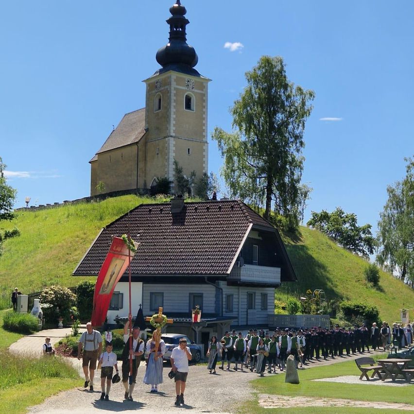 Eine Gruppe von Menschen in traditioneller Kleidung geht auf einem Pfad, mit einer Kirche auf einem Hügel im Hintergrund. Sie tragen eine rote Fahne und ein Kreuz.