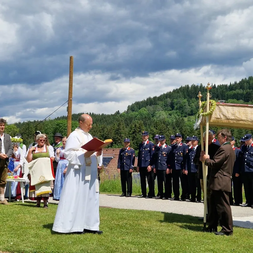 Ein Priester liest aus einem Buch, während eine Reihe von Militärangehörigen in Aufmerksamkeit steht. In der Nähe versammelt sich eine Gruppe von Menschen in traditioneller Kleidung auf einer Grasfläche.