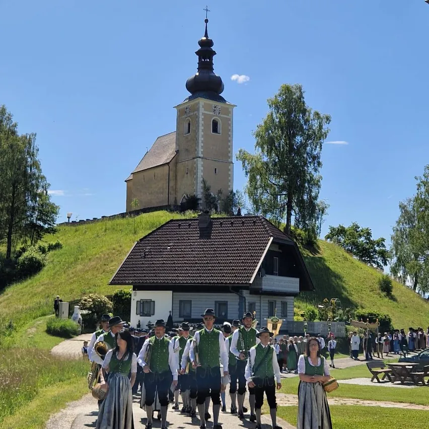 Eine Gruppe von Menschen in traditioneller Kleidung geht an einer Kirche und einem Haus auf einem sonnigen Tag vorbei.