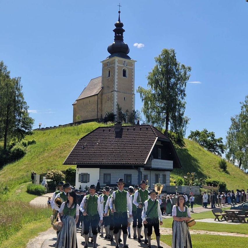 Eine Gruppe von Menschen in traditioneller Kleidung geht an einer Kirche und einem Haus auf einem sonnigen Tag vorbei.