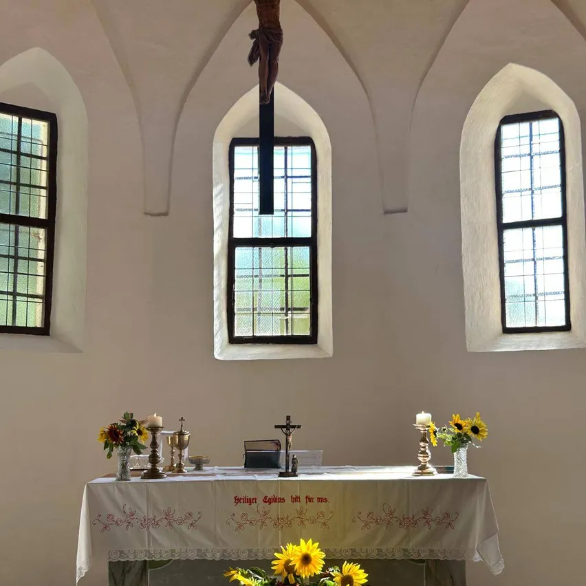 Ein Kirchenaltar mit einem Kreuz darüber, flankiert von bogenförmigen Fenstern, geschmückt mit Blumen, Kerzen und religiösen Texten.