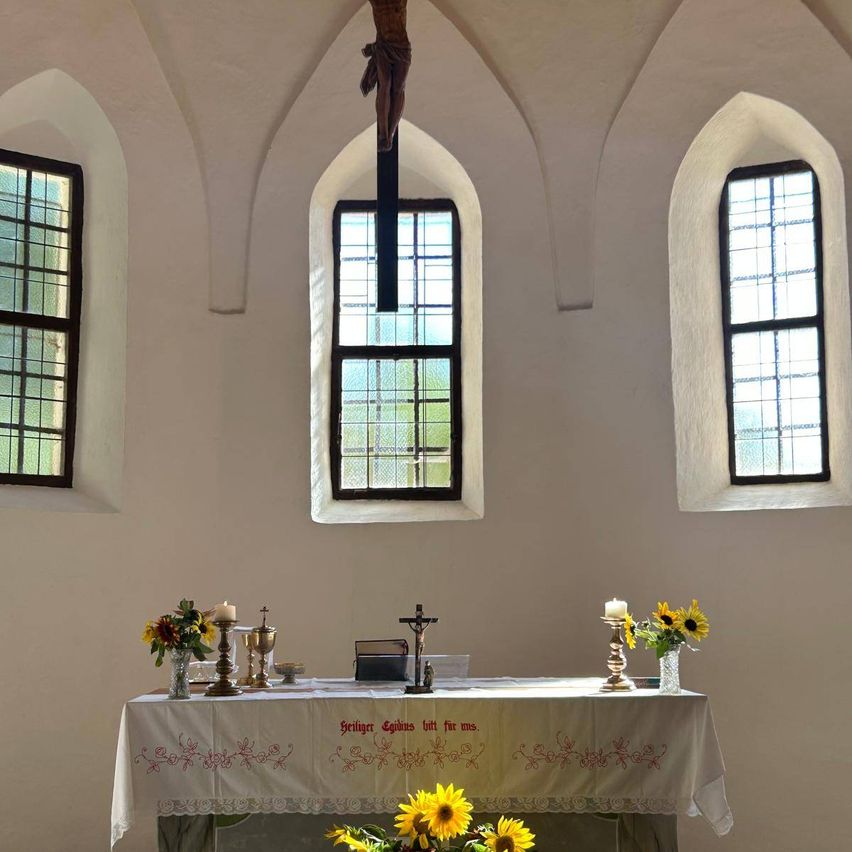 Ein Kirchenaltar mit einem Kreuz darüber, flankiert von bogenförmigen Fenstern, geschmückt mit Blumen, Kerzen und religiösen Texten.