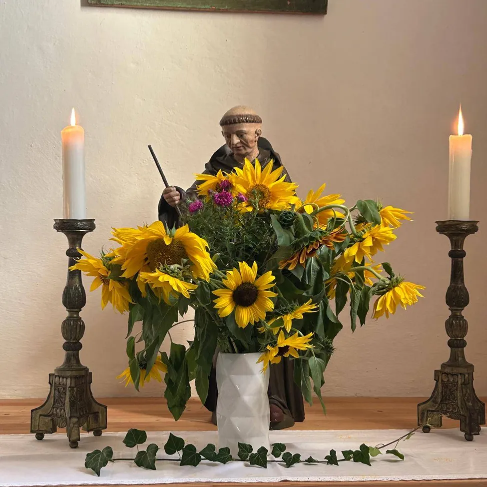 Ein weißer Vase mit Sonnenblumen und anderen Blumen steht zwischen zwei brennenden Kerzen auf einem Tisch. Hinter den Blumen steht eine Statue eines Mönchs mit einem Stock.