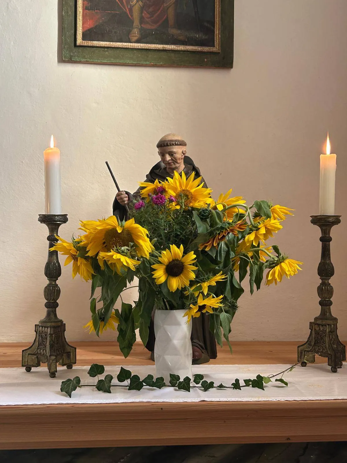 Ein weißer Vase mit Sonnenblumen und anderen Blumen steht zwischen zwei brennenden Kerzen auf einem Tisch. Hinter den Blumen steht eine Statue eines Mönchs mit einem Stock.