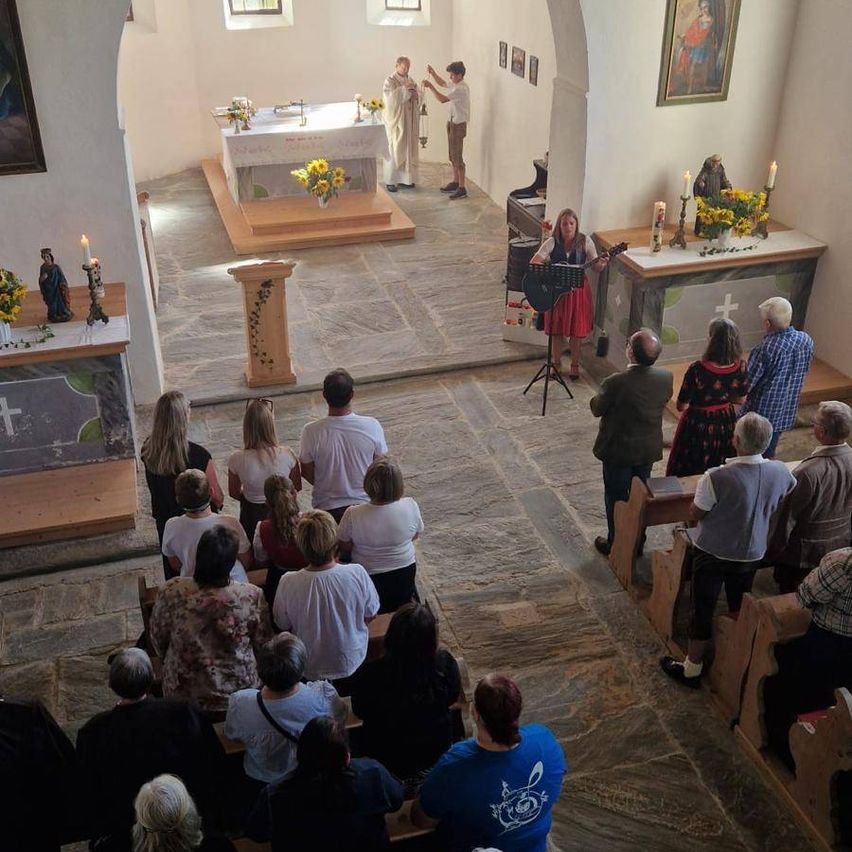 Eine Gruppe von Menschen versammelt sich in einer Kirche, eine Frau spielt Gitarre vor dem Altar. Zwei Priester stehen in der Nähe des Altars, und einige Kerzen sind angezündet.