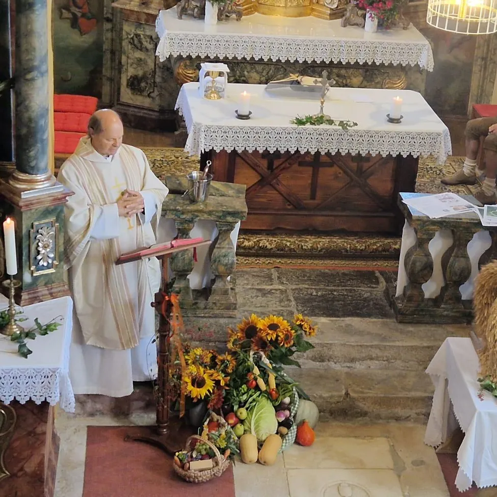 Ein Priester steht an einer Kanzel in einer Kirche, umgeben von Blumen, Kerzen und Gemüse.