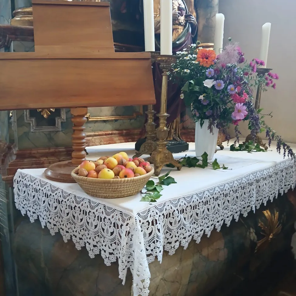 Ein Tisch mit einer weißen Spitzentischdecke, einem Korb mit Äpfeln, einer weißen Vase mit Blumen und einem Kerzenständer mit Kerzen in einer Kirchenumgebung.