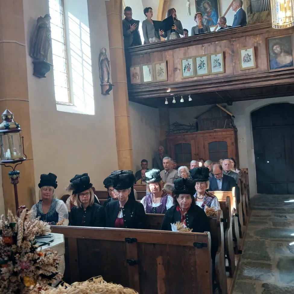 Eine Gruppe von Menschen sitzt auf Kirchenbänken in einer Kirche, wobei einige Personen auf der Empore stehen. Die Kirche hat Holzbänke, einen hölzernen Altar und mehrere gerahmte Bilder an der Wand.