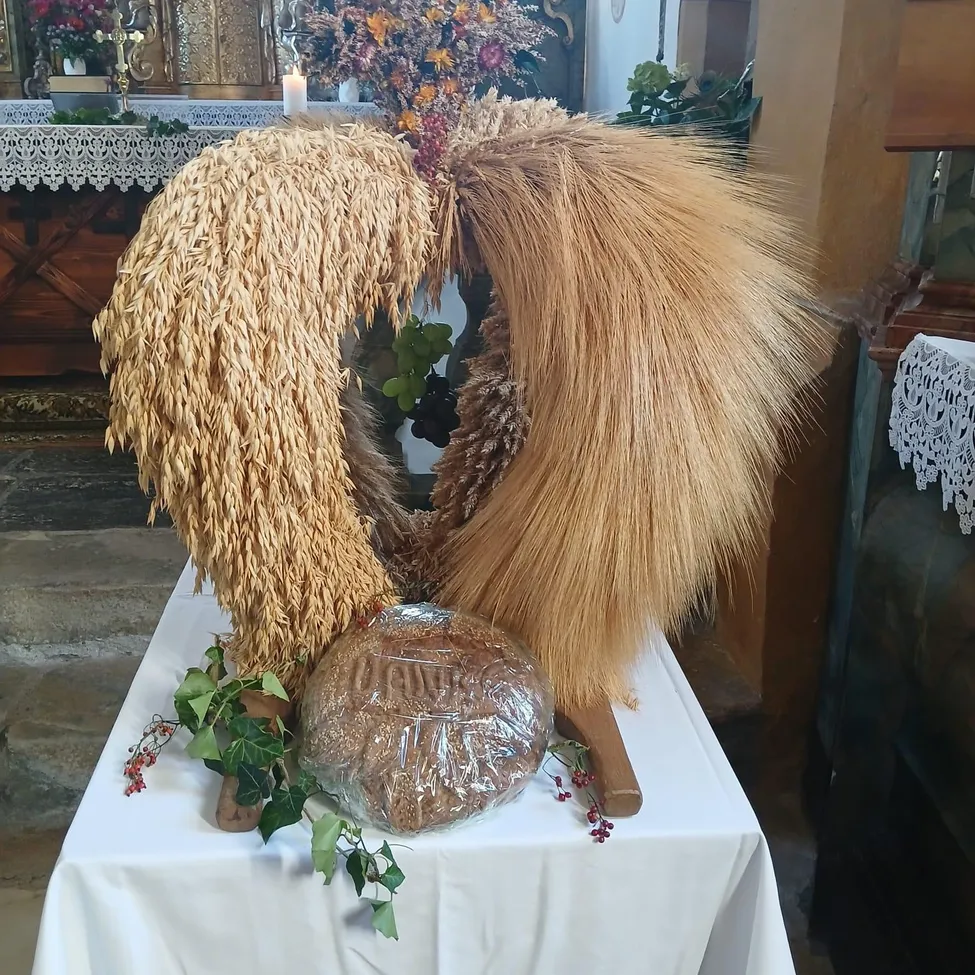 Ein Tisch mit einer weißen Tischdecke ist mit einer Weizenhalm-Girlande geschmückt. Die Girlande liegt auf einem in Plastik eingewickelten Brot. Auf dem Tisch befinden sich auch einige Blätter und eine Kerze.