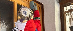 Zwei Personen in traditionellen Kostümen putzen eine Tür. Die Person auf der linken Seite hält ein blaues Tuch, während die Person auf der rechten Seite einen grauen Handschuh trägt. Die Tür hat einen Holzrahmen und Glasscheiben.