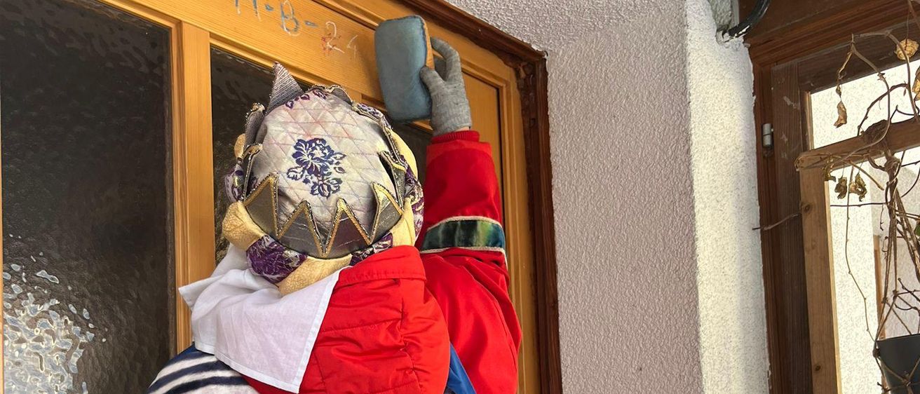 Zwei Personen in traditionellen Kostümen putzen eine Tür. Die Person auf der linken Seite hält ein blaues Tuch, während die Person auf der rechten Seite einen grauen Handschuh trägt. Die Tür hat einen Holzrahmen und Glasscheiben.
