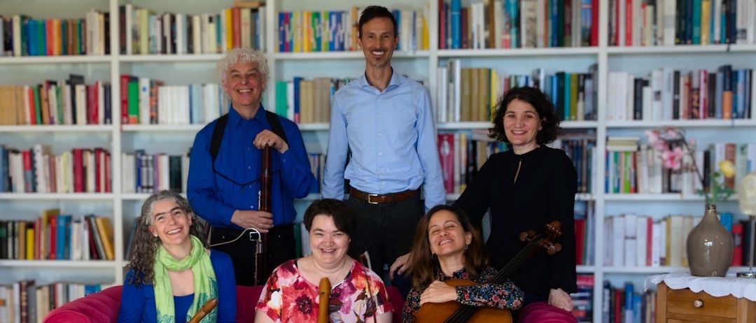 Eine Gruppe von Musikern posiert für ein Foto vor einem Bücherregal, lächelt und hält ihre Instrumente. Sie sitzen auf einem rosa Sofa.