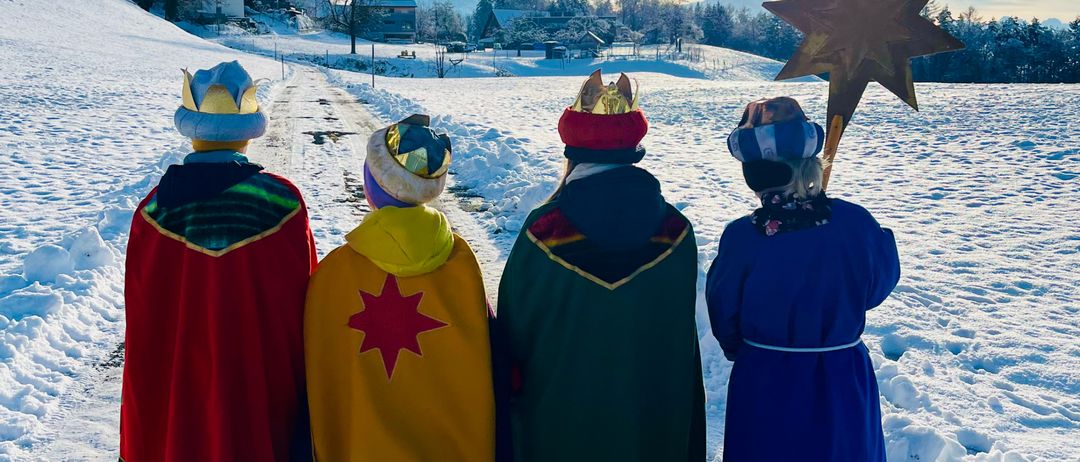 Vier Personen in farbenfrohen Kostümen und Mützen, darunter ein Stern, stehen in einem verschneiten Feld, während die Sonne hinter ihnen untergeht.