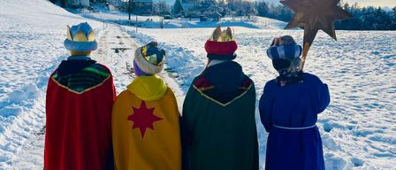 Vier Personen in farbenfrohen Kostümen und Mützen, darunter ein Stern, stehen in einem verschneiten Feld, während die Sonne hinter ihnen untergeht.