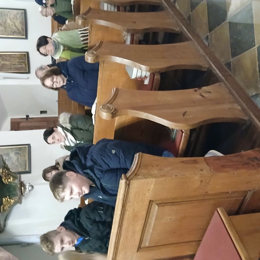 In einer Kirche sitzen mehrere Personen auf hölzernen Kirchenbänken, möglicherweise während eines Gottesdienstes. Die Bänke sind in Reihen angeordnet, und einige Personen tragen Brillen. Die Wände sind mit gerahmten Bildern geschmückt, und der Boden hat ein Schachbrettmuster.